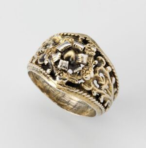  A vintage gold ring with ornate scrollwork and an intricate raised crest or emblem against a light gray background.