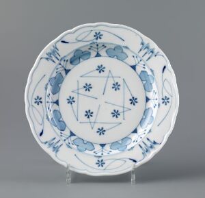  A "Facon Meissen" plate by Gerhard Munthe, made of feldspar porcelain. It features hand-painted underglaze decoration in blue, consisting of a central geometric design surrounded by floral motifs, displayed against a neutral grey background on a clear stand.