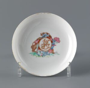  A decorative plate with a scalloped edge featuring a central brown and gold monogram surrounded by a colorful floral motif, including pink flowers and green foliage, displayed on a clear stand against a light grey background. Artist name and title are unknown.