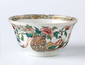  An ornate white porcelain bowl with a wide opening and gold trim, featuring intricate hand-painted floral patterns and decorative vases, showcasing hues of pink, green, blue, and touches of gold.