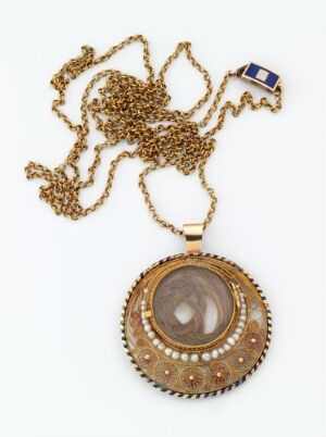  An intricately designed vintage gold-toned pendant with a central transparent stone and pearl-like adornments, attached to a gold chain with a small blue rectangular detail, against an off-white background.