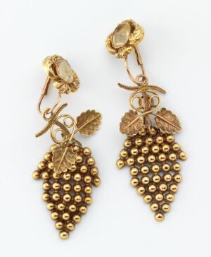  A pair of gold-toned earrings in the shape of grape clusters with detailed leaves and stems, featuring numerous small beads to represent grapes, reflecting light with a polished finish. The artist's name and title are unknown.