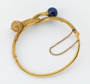  An antique gold earring in a 'D' shape with a twisted, rope-like textured clasp and a dangling smaller gold hoop. A single dark blue bead, likely lapis lazili, is set opposite the clasp end against a neutral background.