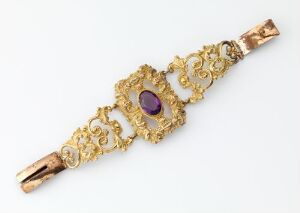  A detailed piece of gold jewelry with baroque patterns and a central, oval-shaped purple gemstone—artist and title unknown.