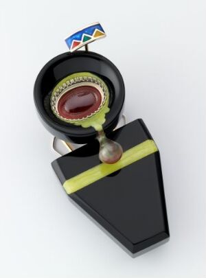  A brooch designed by Morten Haugmo titled "Føler-fjell," featuring a geometric and abstract design with a small rectangular element with primary colors at the top, and a large black triangular body with a neon yellow stripe and a circular feature with a maroon cabochon in the center. A shiny sphere hangs off the bottom, adding to