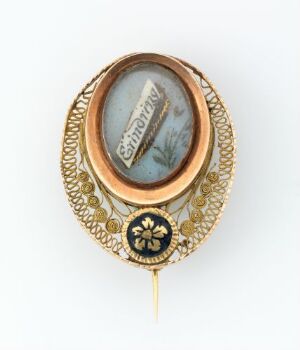  A vintage oval brooch featuring a domed glass centerpiece with a protected miniature artwork or message, surrounded by a detailed golden filigree frame, and a small circular emblem with a golden flower on a dark background at the bottom.