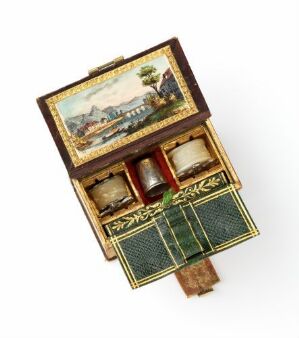  An antique, miniature wooden chest of drawers, with the top lid open showing a small painted rural scene, revealing four small drawers with golden trim and handles.