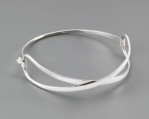  "Sløyfe," a hand-forged sterling silver bracelet by Tone Vigeland, features an elegant and minimalistic overlapping ribbon design with a reflective, silver metallic finish against a neutral gray background.