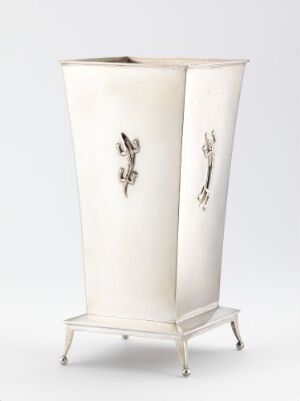  An elegantly tapered pedestal stand in a light ivory color with decorative metallic handles on the sides, supported by a simple metallic frame and legs against a plain white background. Artist name and title are unknown.