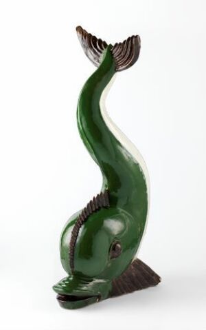  A stylized, glossy emerald green sculpture of a fish arching backwards, with dark scale details and contrasting brownish-grey fins and base.