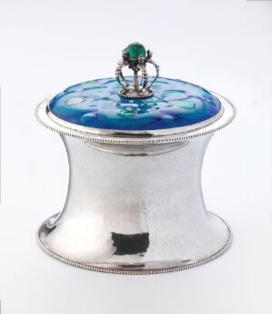  An ornate silver-colored container with a reflective surface and a blue enameled lid adorned with a green gem atop an intricate metal framework, against a white background.