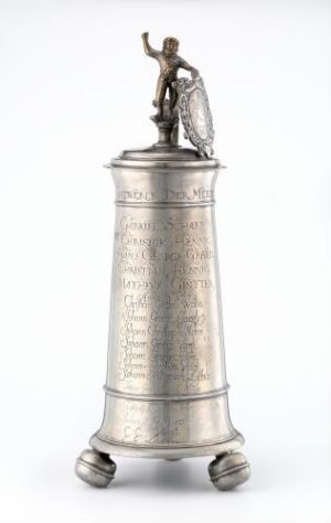  A 'Drikkekanne' by an unidentified artist, featuring a tall, cylindrical pewter pitcher with engraved decoration, set on three rounded feet, and topped with a small, cast brass figurine on the lid. The color varies from silvery-grey of the pewter to the golden hue of the brass figure.