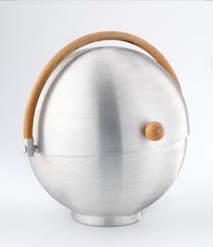  An "Informal Serving Accessories" spherical container by artist Russel Wright, made from brushed aluminium with a curved wooden handle on top and a round wooden knob on the front, set against a soft grey background.