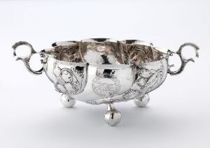  A silver bowl titled "Skål" by Søren Joensen, with a gleaming finish and embossed decorative patterns. The bowl features elaborate handles and a stabilizing base, all exhibiting intricate chiseled and engraved designs.