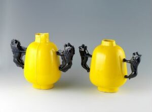  "Elle melle" by Heidi Bjørgan, featuring two vibrant yellow porcelain vessels with black human-like hands as handles, displayed against a light-grey gradient background.