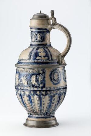  An antique ceramic jug with cobalt blue and white designs, featuring intricate patterns and oval medallions. The jug has a hinged lid on the spout and a robust handle, set against a neutral background.