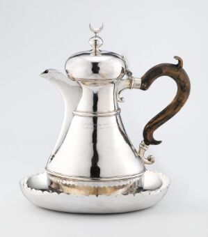  A silver coffee pot with a carved wooden handle and accompanying saucer designed by Jacob Nilsen, named "Kaffekanne og underfat," displayed against a light gray background. The pot and saucer show polished surfaces with some punched decoration, conveying a sense of traditional elegance.