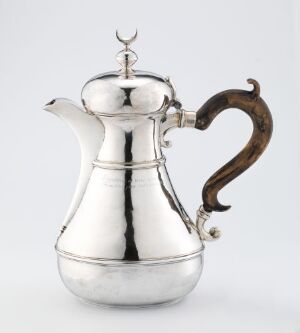  An antique silver coffee pot with a shiny surface and a dark, curved handle against a light background.