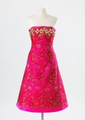  A strapless, A-line evening gown designed by Ove Harder Finseth, titled "Orientalsk blomsterhave." The vibrant pink silk fabric is embroidered with a floral pattern and embellished with gold threads, glass beads, and appliqued gold flowers. The gown is displayed on a mannequin against