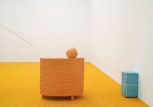  An art installation by Anders Herwald Ruhwald titled "You in between" features a large terracotta glazed stoneware block with a spherical object on top in the foreground, and a smaller turquoise block to the right, all on a mustard-yellow floor against a white wall.