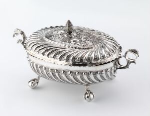  An elaborately decorated oval silver tureen with ribbed patterning and scrolled handles, displayed against a plain background.