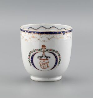  A porcelain teacup with decorative blue, gold, and red detailing around the rim and a stylized emblem in the center featuring a vase with floral design, set against a plain white background with a reflective surface.