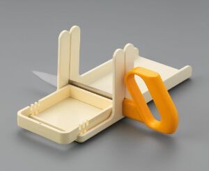  A household kitchen gadget designed by AB Gustavbergs Fabriker, featuring a cream-colored propylene plastic base with vertical arms and a stainless steel blade, and an orange ergonomic handle. The object seems intended for food preparation, possibly slicing, and is displayed against a soft gray backdrop.