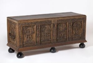  An antique oak chest named "Kiste" with detailed carved relief decoration on the front, showing several figures within framed sections, supported by rounded bun feet. The chest displays a dark brown patina indicative of aged oak, artist unidentified.