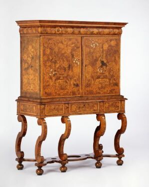  An antique wooden cabinet with ornate inlays on its doors, placed on a decorative stand with S-shaped legs, set against a plain background.