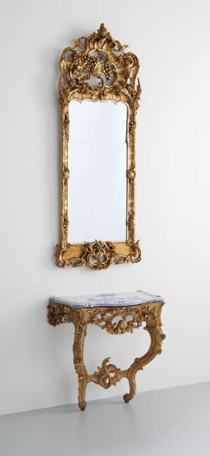  An unidentified artist's "Konsollbord," featuring an intricately carved and gilded birch mirror frame above a matching console table with a white-glazed faience tabletop decorated with hand-painted designs, set against a plain wall.