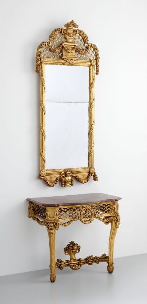  An ornate gold-framed mirror with a crest-like top and a matching console table with a white marble top, both featuring elaborate acanthus leaf carvings against a light gray wall. Artist name and title are unknown.