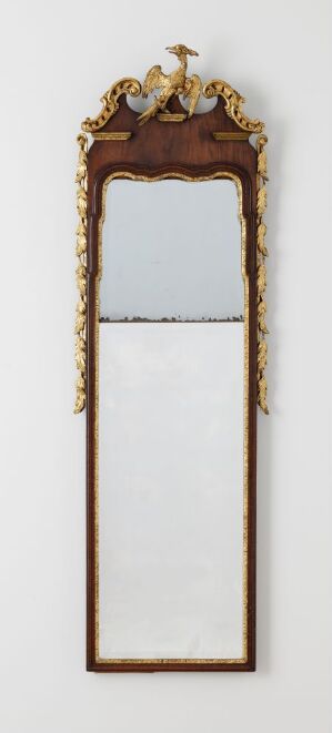  An ornate, baroque-style tall vertical mirror with a decorative gold frame, featuring an intricate crest at the top and curled leaf motifs along the sides, reflecting a neutral light grey background.