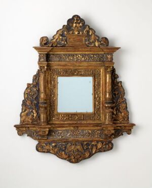  An antique gilded wooden mirror mounted on a light-colored wall, featuring baroque style carvings including cherub figures and ornate scrollwork, with a small centered mirror glass panel.