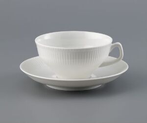  A white feldspar porcelain cup and saucer set with linear relief decoration designed by Tias Eckhoff on a gray background. The set exemplifies a minimalist and elegant design with its smooth, glossy surface and tactile linear grooves.