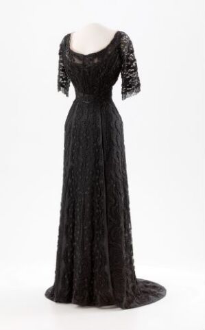  A black evening gown with a delicate texture and three-quarter-length sleeves on a mannequin against a neutral background. The dress features a fitted bodice, scoop neckline, and a floor-length skirt that pools gently at the base.