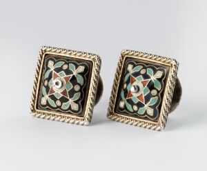  A pair of ornate square cufflinks with a twisted rope-like gold-tone border and a mosaic-style inlay of white, black, green, red, and gold, centered with a pearl-white motif, on a light grey background. Artistname and title are unknown.