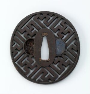  A circular dark metallic object with a symmetrical maze-like cut-out pattern, possibly a Japanese sword hand guard (tsuba), on a light background. Artist name and title are unknown.