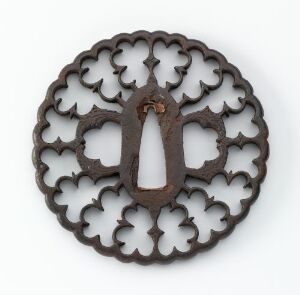  A circular, dark oxidized iron Kamakura-tsuba with a delicate, openwork floral design, created by the Owari School, against a plain background. The iron handguard shows a complex pattern of interlacing shapes, carved and pierced through the metal, with small, barely visible inlays of shakudo adding a hint of shimmer.