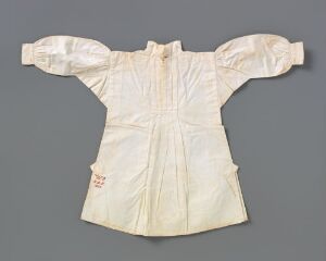  An antique creamy white infant's christening gown with delicate embroidery on the collar and sleeves, pleated detail on the body, and a small emblem near the waist, displayed against a plain gray background.