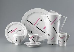  A collection of modern white porcelain tableware with abstract black and pink geometric designs, designed by Poul Jensen, displayed against a grey background. The set includes plates, a teapot, cups, and saucers, all showcasing a clean and elegant appearance with strong contrast between the elements.