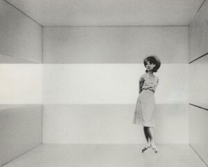 
 A black and white photograph of a minimalist architectural interior featuring a broad horizontal band of light across the middle of the walls. A woman stands off-center in the room, dressed in light attire, with a relaxed posture under the soft illumination, emphasizing the stark, geometric simplicity of the space.