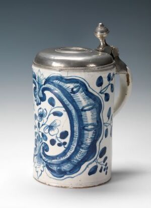  A traditional white and cobalt blue ceramic beer stein with a metal lid, featuring intricate hand-painted designs and a sturdy handle, set against a neutral light grey background.