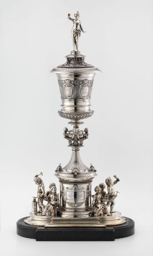  An elaborate silver trophy with a tiered design featuring a figurine on top, ornamental figures around the base, and intricate decorative details throughout, set against a light background.