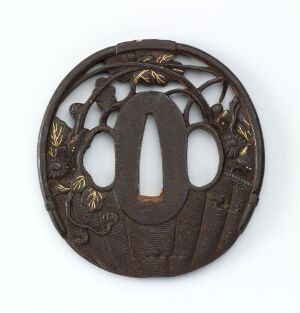  An intricate, round, dark brown metallic tsuba, featuring a central elliptical opening flanked by two small circles, with ornate openwork and gold-leaf accents of branches and leaves. Artist name and title are unknown.