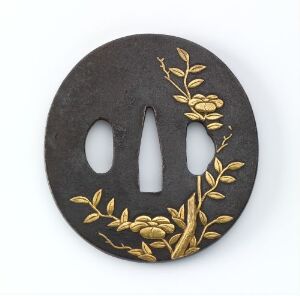  A circular dark grey or black metal object with two vertical slits and a teardrop-shaped cutout, adorned with a golden embossed branch with leaves motif on the right side, suggesting an item of decorative or functional accessory.