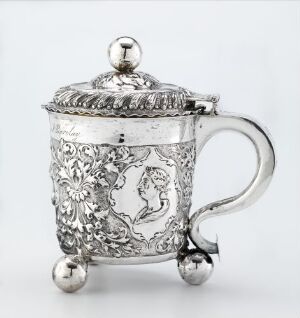  "Drikke kanne," a silver drinking pitcher with embossed and chased floral and figural decoration, resting on three spherical feet, with a curved handle and ornately patterned lid with a finial, crafted by Johannes Johansen d.e. Reimers. The intricate design reflects light with a sophisticated interplay of polished and matte finishes.