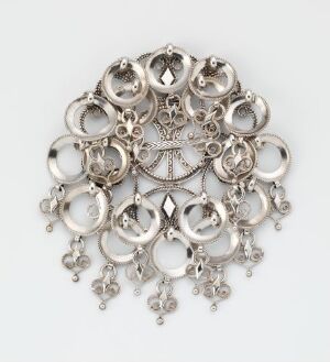  A detailed, silver, circular piece of jewelry with concentric rings of decorative filigree work and dangling elements on a muted gray background. Artist name and title are unknown.