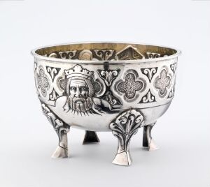  A silver ornamental bowl with a metallic sheen featuring dark embossed or engraved patterns, including fleur-de-lis motifs and portrait faces, resting on three leaf-shaped feet.