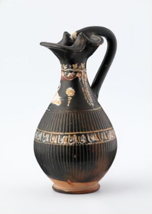  An ancient-styled ceramic jug with a black glaze and decorative frieze in the center, featuring a sculpted animal head at the neck and a warm terracotta base, set against a neutral background.