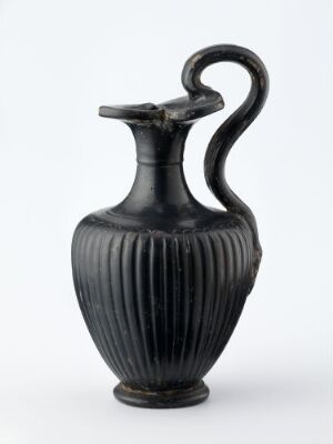  An ancient ceramic jug with vertical ribbed texture, matte black in color, featuring a gracefully curved handle and a narrow spouted mouth against a light grey background.